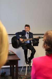 Jacek Mianowski siedzący na stołku, śpiewający i grający na gitarze